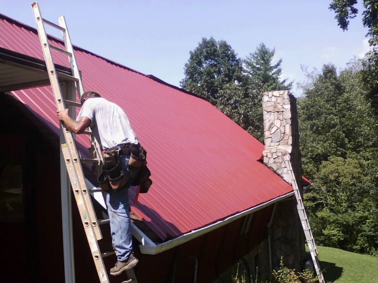 Roofer on a ladder beside a red metal roof and stone chimney — Soffit & Fascia Repair by Roofing Brothers in Ashley Falls